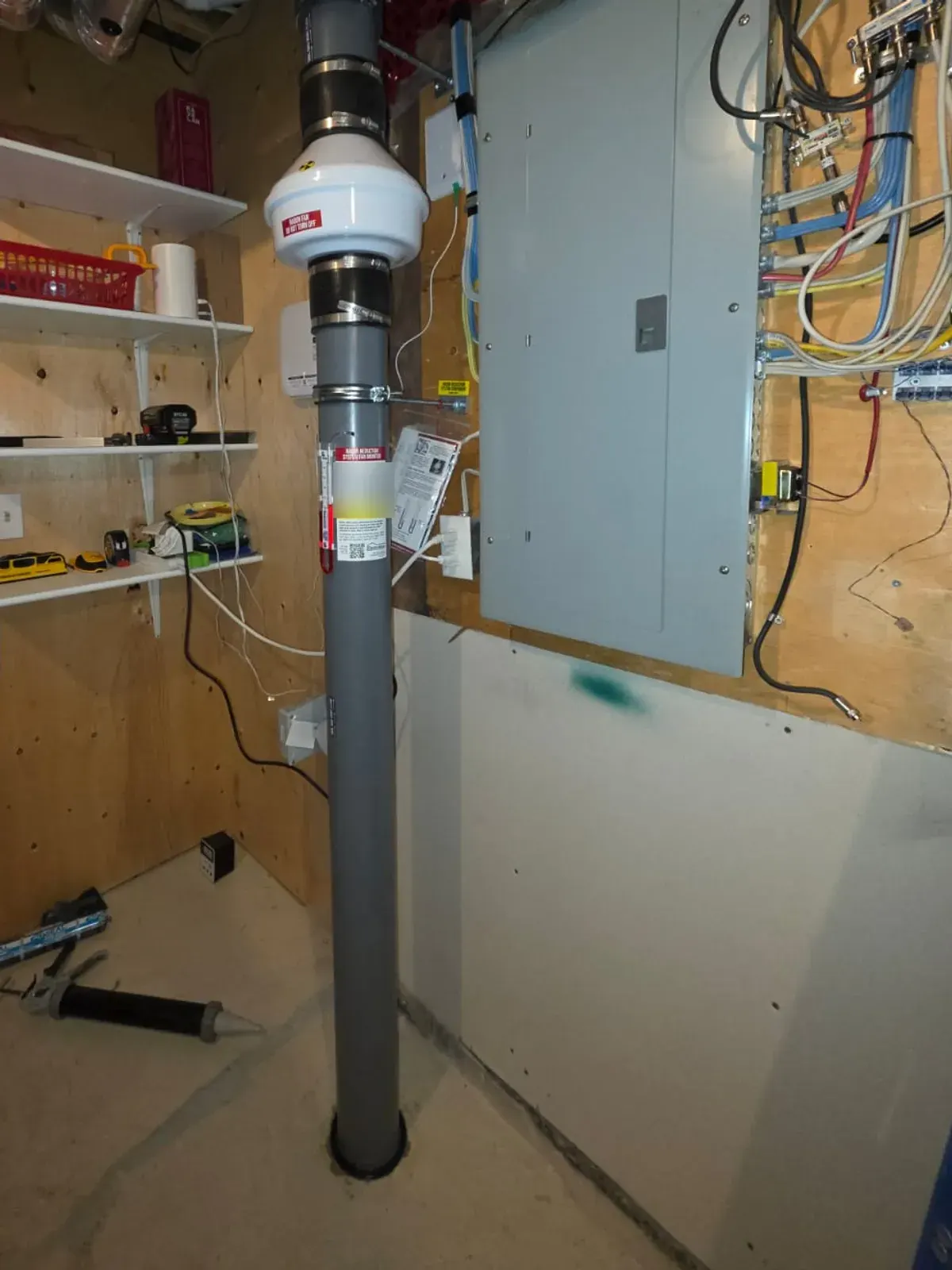 Radon fan and vent pipe installed next to the electrical panel in a basement storage room